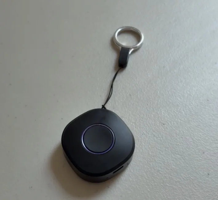 Photo of the StepSafe tracker, a small black rounded device with a ring on its face, on a lanyard loop on a table.